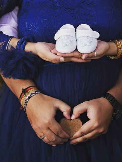 A creative close-up shot symbolizing teamwork and love. The mom-to-be holds the baby shoes while her partner makes a heart shape with his hands around her bump.