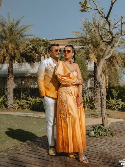 A stylish shot of the couple posing in their coordinated yellow and white outfits against the lush greenery of the venue.