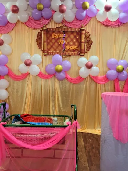 A sweet naming ceremony setup for a baby girl, using purple, pink, and white balloons to create flower shapes. The simple cradle is draped in pink netting.