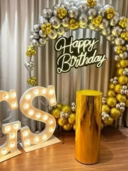 A personalized birthday setup with large marquee-light initials. The backdrop features a ring of gold and silver balloons, a custom 'Happy Birthday' sign, and a golden plinth for the cake.