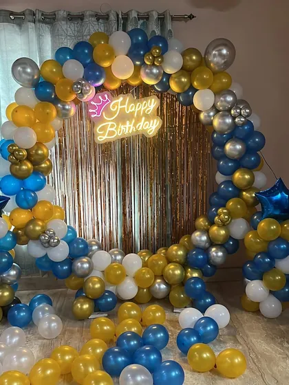 A vibrant birthday setup with a balloon ring of blue, yellow, white, and silver balloons, complete with a foil curtain and neon sign.