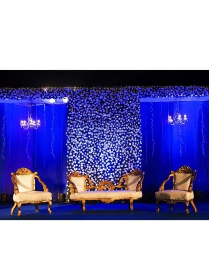 A striking reception stage with a wall of white flowers set against deep blue lighting. The contrast creates a dramatic and modern look, complemented by ornate gold and white seating for a touch of royalty.