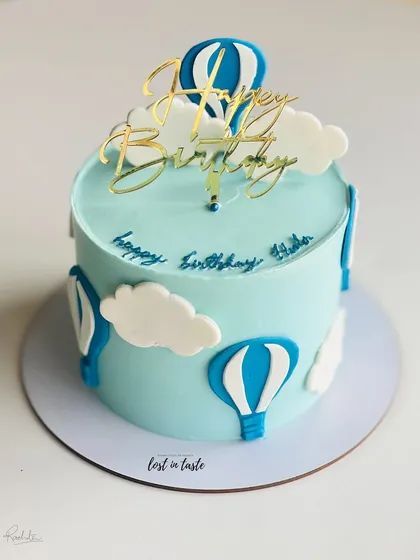 A whimsical hot air balloon themed cake. The light blue buttercream sky, fluffy clouds, and a golden "Happy Birthday" topper make it a delightful choice for a child's celebration.