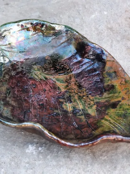 The magic of Raku firing. This leaf-shaped platter started as plain clay and was transformed by fire and smoke into this iridescent, unpredictable piece of art. This is what happens when you let the process have a say.