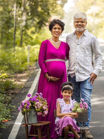 A classic family portrait taken on a quiet road surrounded by nature. The older daughter is seated with a bouquet, and the whole family is smiling, full of joy.