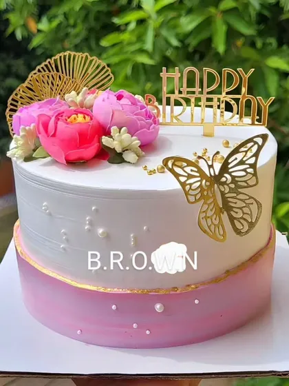 A beautiful and elegant birthday cake. The design combines a pink and white ombre effect with fresh pink flowers, a gold butterfly, and a "Happy Birthday" topper.