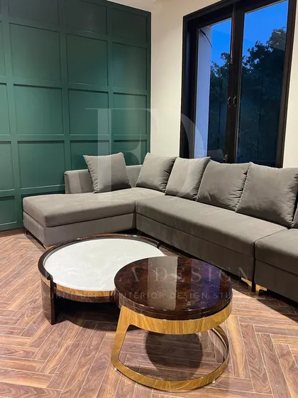 A grey L-shaped sectional set against a dark green paneled wall. The herringbone wood floor and nested marble coffee tables with brass accents complete this moody and sophisticated living room.