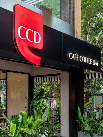 A close-up of the iconic red CCD logo, which glows invitingly. The surrounding greenery softens the modern architecture and connects the cafe to its garden city setting.