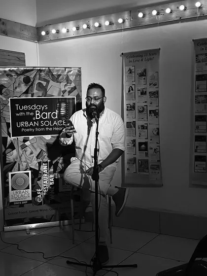 A poet, seated and comfortable, shares his work with an engaged audience. The monochrome tones emphasize the emotion of the performance.