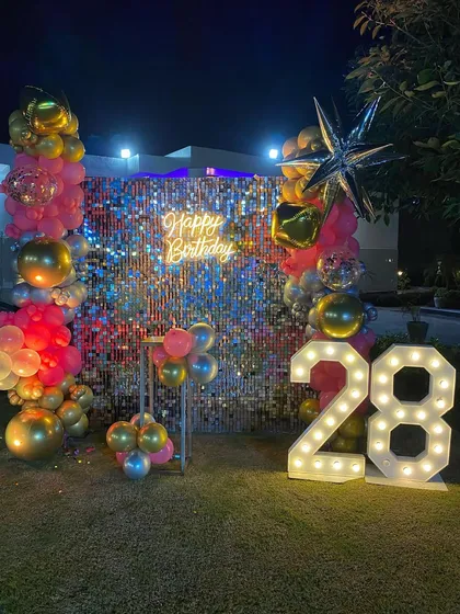 An outdoor shimmer wall setup for a 28th birthday. The pink, gold, and silver balloons, along with the neon sign and marquee numbers, create a festive party vibe.