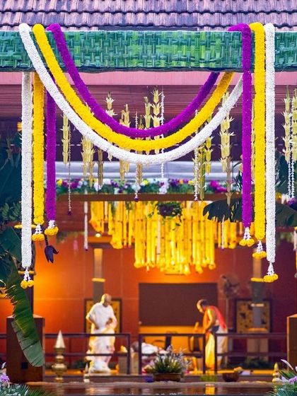 The main wedding arch for a traditional South Indian Muhurtham, constructed with woven coconut leaves and adorned with garlands of yellow, purple, and white flowers.