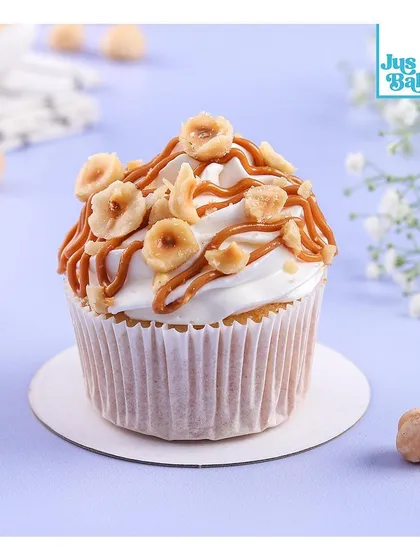 A close-up of our Biscoff Hazelnut cupcake. This gourmet cupcake has a vanilla base, is topped with a light cream, drizzled with caramel, and generously garnished with toasted hazelnuts.