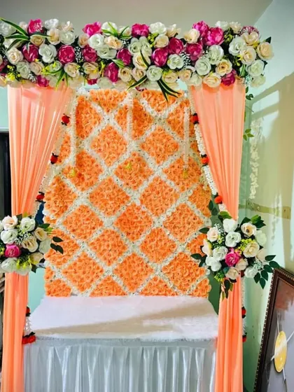 A full wall backdrop created with a lattice of orange and white artificial flowers. This textured and vibrant background is framed by peach-colored drapes, offering a unique and eye-catching setting for Bappa.