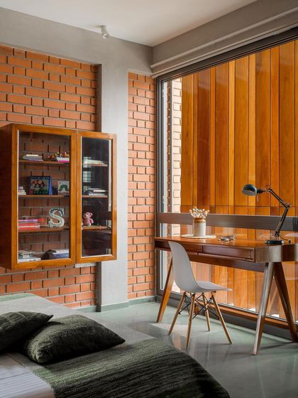 The bedroom's study nook is set against an exposed brick wall, with a large window screened by the wooden louvers. This creates a warm, private space for work or reading, with beautifully filtered natural light.