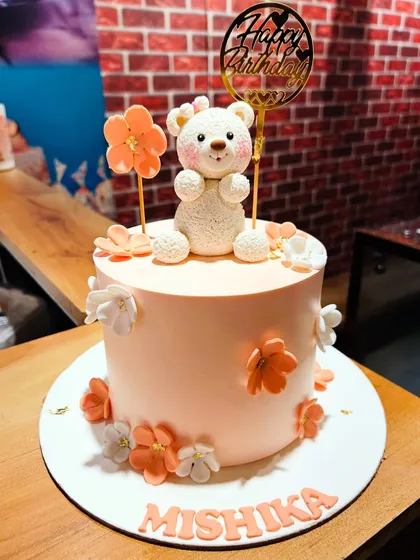 A sweet peach-colored cake with a fluffy teddy bear topper and delicate flowers.