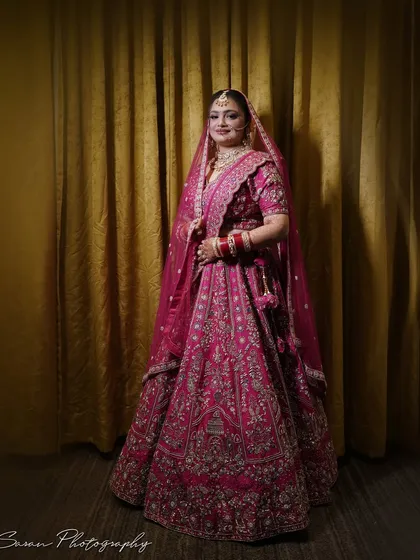 A classic full-length portrait of the bride in her vibrant pink lehenga against a golden curtain.