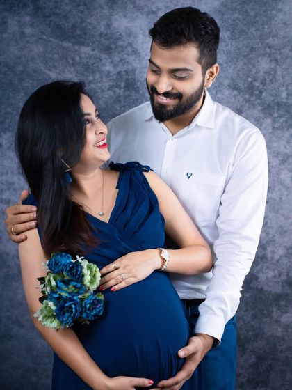 A classic and happy couple's portrait. The mom-to-be holds a bouquet of blue flowers while her partner stands with her, both smiling at the camera.