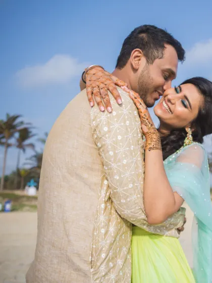 A joyful embrace on a sunny beach. The bride's henna-adorned hands and the couple's radiant smiles capture a moment of pure happiness with palm trees in the background.