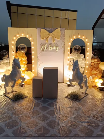 A full view of the stunning horse-themed "Oh Baby" shower at dusk. The warm glow from the lights on the arches and the neon sign creates a magical and inviting atmosphere, with two horse statues framing the beautiful setup.