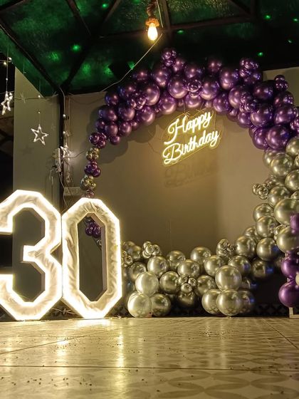 A dramatic outdoor setup for a 30th birthday. This features a half-arch of metallic purple and silver balloons against the night sky, a neon sign, and giant illuminated marquee numbers.