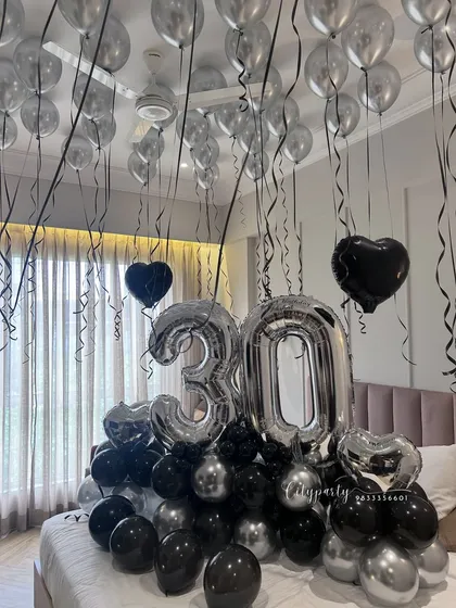 This is an example of our surprise birthday room decoration service for a 30th birthday. The setup fills the room with helium balloons on the ceiling and features a large balloon bouquet on the bed with silver foil numbers and black and silver accent balloons.