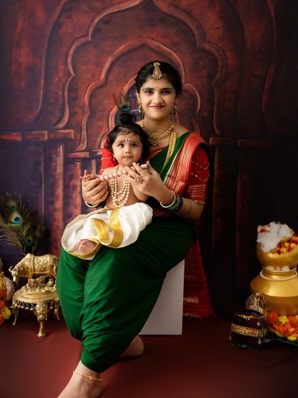 A mother dressed in a traditional green saree holds her baby, who is dressed as Krishna. This portrait captures the beautiful bond between mother and child during the festive season.