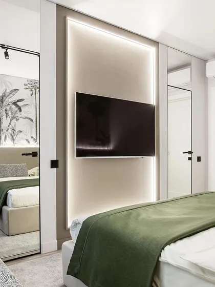 A modern bedroom design featuring a TV wall with integrated backlighting. The room includes a full-length mirror that reflects the bed and its tropical-themed accent wall, making the space feel larger.