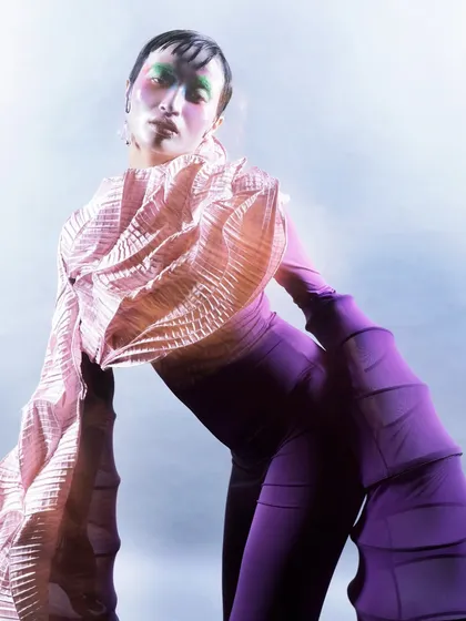 A detailed shot focusing on the textured, pleated fabric and the model's expressive pose. The lighting creates a soft, ethereal glow that contrasts with the structured garment.