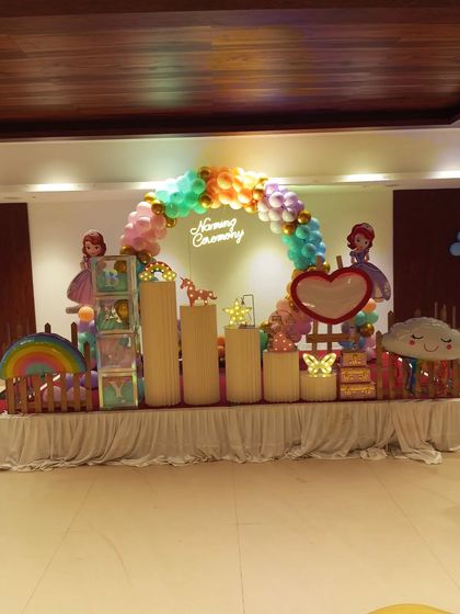 A rainbow princess theme for a Naming Ceremony. This setup features cutouts of Princess Sofia, a rainbow, a unicorn, and a multi-colored balloon arch over a 'Naming Ceremony' neon sign.