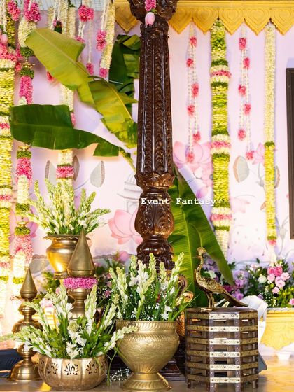 Another detail shot showing the rich textures of the decor. A carved wooden pillar stands next to banana leaves and brass pots, with hanging floral garlands adding color and fragrance.