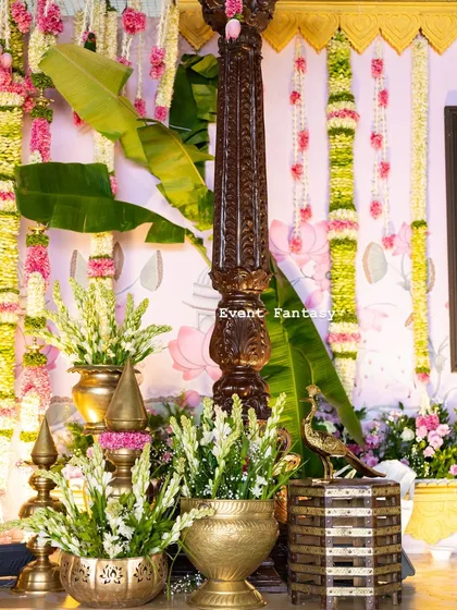 Another detail shot showing the rich textures of the decor. A carved wooden pillar stands next to banana leaves and brass pots, with hanging floral garlands adding color and fragrance.