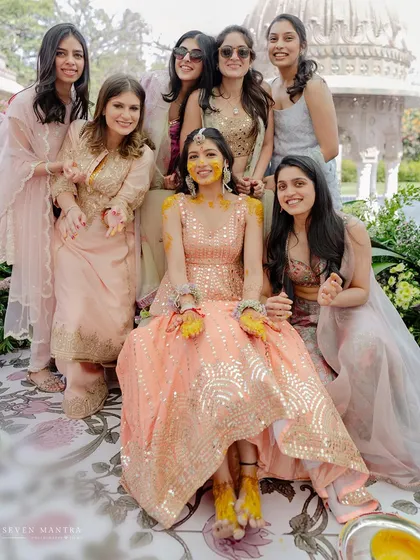 A bride surrounded by her best girls. The coordinated pastel outfits of the bridesmaids complement the bride's shimmering peach lehenga, creating a beautifully styled group portrait full of smiles.