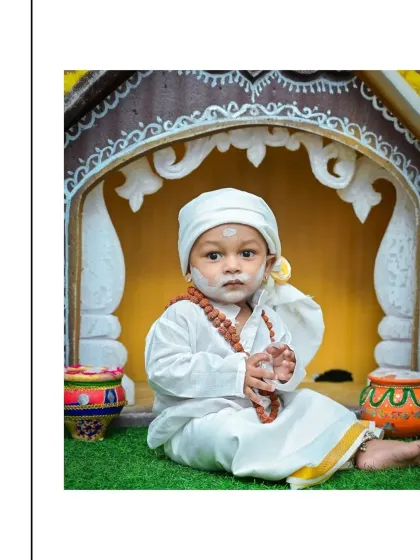 A baby boy dressed as a young Sai Baba, sitting peacefully in front of a miniature temple. The white robes, head covering, and prayer beads complete this serene and spiritual look.