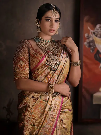 A model in a regal pose, wearing a heavily embroidered blouse with a gold and beige patterned saree. The full-sleeve embroidery adds a layer of luxury to the ensemble.