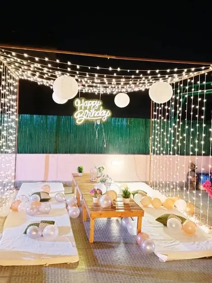A beautiful terrace setup for a birthday. This decoration features a canopy of fairy lights, low seating, and a "Happy Birthday" neon sign, creating a warm and inviting atmosphere.