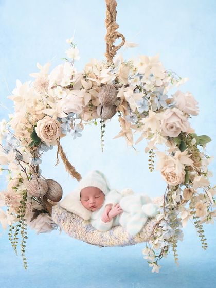 A baby sleeps in a hanging wreath decorated with white and blue flowers against a painted sky backdrop, creating a dreamy, floating-on-a-cloud effect.