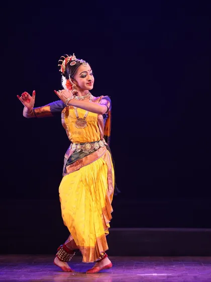 A solo dancer in a graceful pose, her yellow costume radiant against the dark stage, from the Sadhana festival.