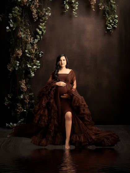 A moody and romantic fine art portrait. The mother-to-be stands in a dark, rich brown gown against a deep-toned background with hanging foliage and a subtle water reflection.