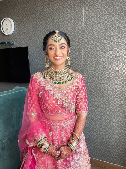 Radiating her inner glow on her wedding day. This look features a classic bridal bun and dewy, natural makeup that complements her beautiful pink lehenga and emerald jewelry.