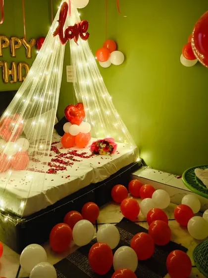 This birthday setup features a fairy light canopy over the bed, with red and white balloons scattered on the floor and a "love" foil balloon adding a romantic touch.