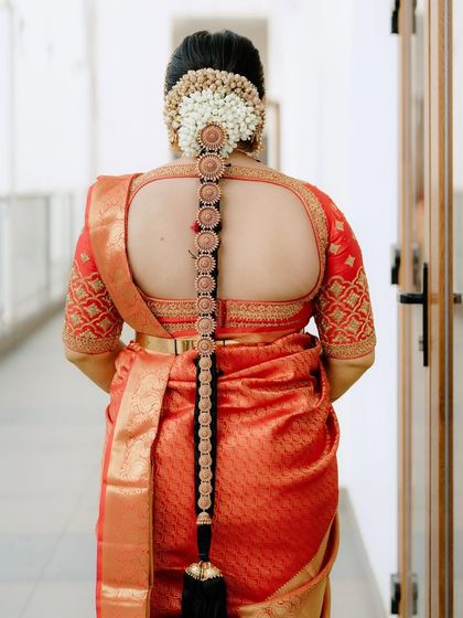 The back view of a bride in an orange Kanchivaram, showing the elegant cut-out design of her blouse and traditional South Indian hair adornments.