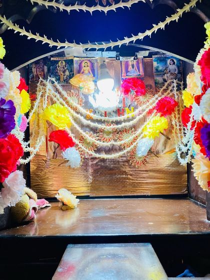 A close-up of a home mandir decorated with bright, colorful artificial flowers and delicate pearl strings. The light inside highlights the decorations and the deities.