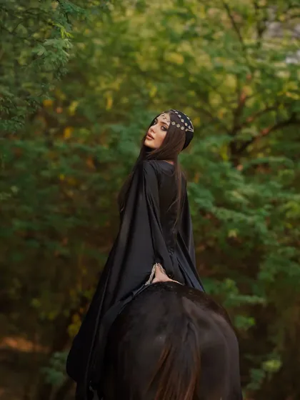 A model in a flowing black robe on horseback, creating a striking silhouette against the green foliage. The natural environment enhances the drama of the shot.