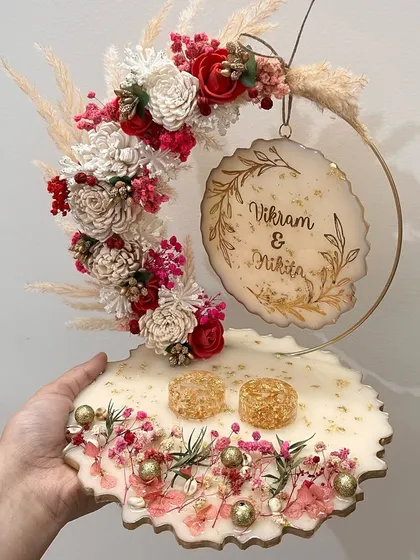 This custom resin ring platter is one of my absolute favorites to create. The design features a beautiful floral hoop with preserved red and white flowers, pampas grass, and delicate gold details, all mounted on a geode-shaped base. It's the perfect statement piece for your engagement ceremony.