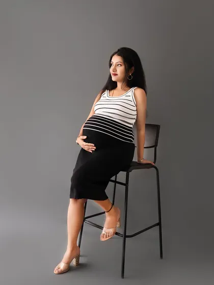 A confident solo portrait of a mom-to-be in a modern striped outfit, seated on a high chair against a clean grey background.
