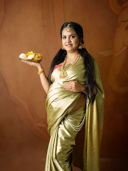 A portrait of a mom-to-be in a traditional green saree, holding a plate of offerings, with a painted Ganesha in the background for blessings.