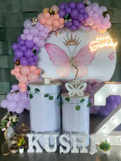 A butterfly theme for a second birthday celebration. The decor includes a marquee number '2', a neon "Happy Birthday" sign, and personalized name blocks in front of the pastel balloon backdrop.