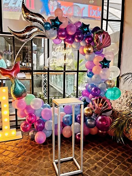 A magical mermaid-themed first birthday decoration. This setup features a balloon arch with large mermaid tail and seashell foil balloons, a "Happy Birthday" neon sign, and a light-up number "1".