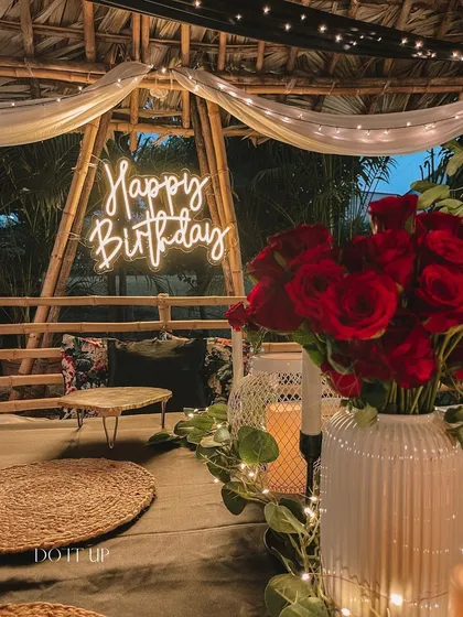 A cozy and rustic birthday setup. The 'Happy Birthday' neon sign adds a modern touch to the bamboo gazebo, while soft drapes and floor seating create a relaxed and intimate atmosphere.