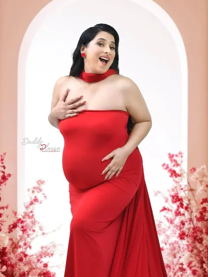 A joyful and expressive portrait of a mom-to-be in a bright red strapless gown. The simple arched backdrop and floral accents keep the focus on her happy expression.